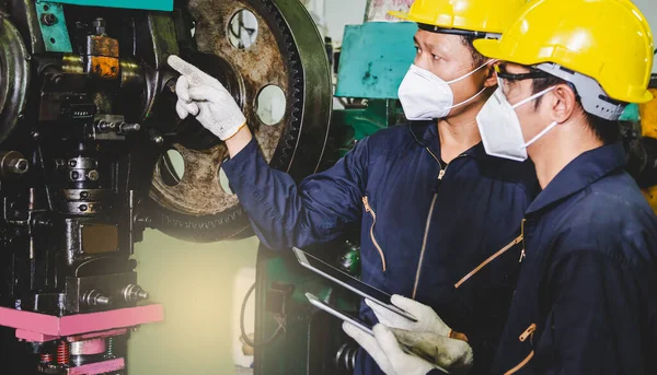 Metal fabrikası mühendisleri, kasketli, maskeli ve eldivenli. Tablet bilgisayarlarını endüstriyel bir tesisin içinde tornalı metal parçaları üretmek için tutuyorlar..