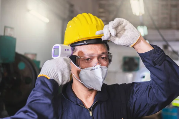 A male worker wearing gloves and a scanning temperature worker that is wearing a face mask and hard hat before entry inside the industrial facility to protect the virus COVID 19 for the worker in the factory.