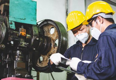 Metal fabrikası mühendisleri, kasketli, maskeli ve eldivenli. Tablet bilgisayarlarını endüstriyel bir tesisin içinde tornalı metal parçaları üretmek için tutuyorlar..