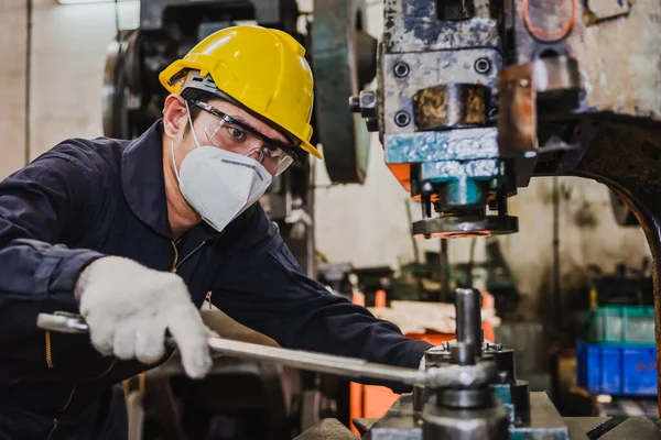 Asian factory engineer in uniform, hard hat, gloves, and a face mask is working hard in the factory with a lathe.