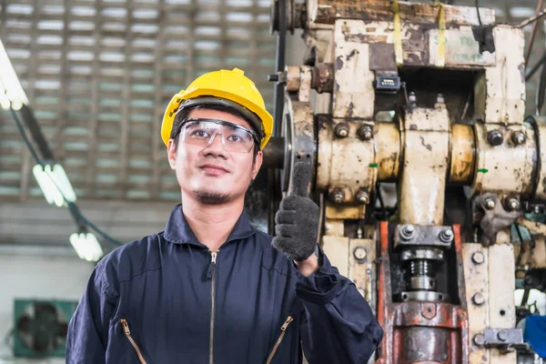 Üniformalı Asyalı bir fabrika işçisi ve sarı bir şapka fabrikada çalışırken ayakta duruyor ve başparmağını kaldırıyor..