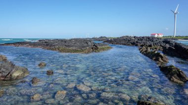 Körfezin güzel deniz manzarası, kristal berrak su ve lav resifleri ön planda ve Güney Kore adası Jeju 'da rüzgar jeneratörleri.