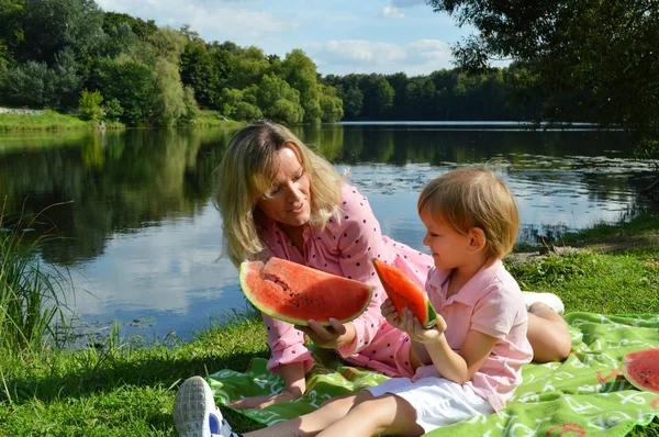 Niño pequeño con madre en la orilla del río comer sandía . — Foto de Stock