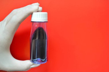 Beyaz kauçuk eldiven içinde kırmızı zemin üzerinde sıvı bulunan plastik şeffaf bir şişe tutar.