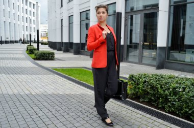 Modern ofisin arka planında güzel bir iş kadını. Siyah tulumlu ve kırmızı ceketli bir kız iş merkezinin yanında yürüyor.