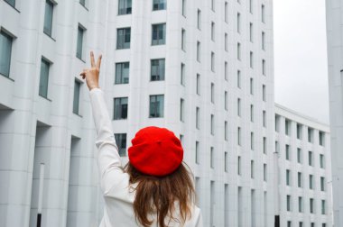 Modern ofisin arka planında güzel bir iş kadını. Siyah saçlı, beyaz ceketli, kırmızı bereli elini iş merkezinin yakınına kaldırmış bir kız. Fotoğraf arkadan.