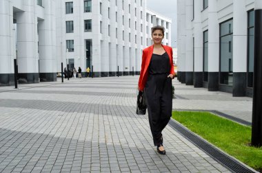 Modern ofisin arka planında güzel bir iş kadını. Siyah tulumlu ve kırmızı ceketli bir kız iş merkezinin yanında yürüyor.