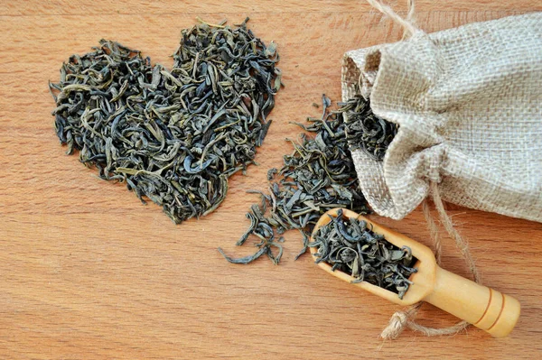 Heart made from loose leaf tea, jute bag and wooden spoon on wooden background