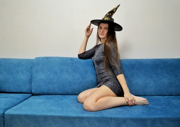 Uma jovem se senta em um vestido brilhante no sofá azul com um chapéu de bruxa na cabeça — Fotografia de Stock