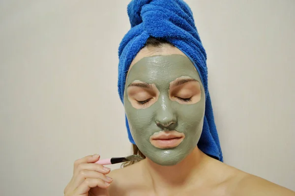 A woman with a brush applies a clay mask to her face with a blue towel on her head