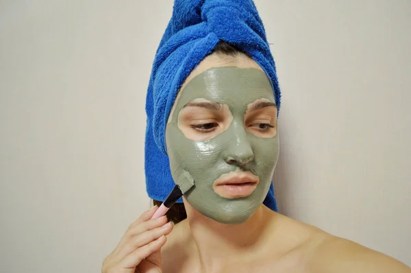 一个拿着刷子的女人用粘土面罩盖住她的脸 头上还戴着一条蓝色毛巾 — 图库照片