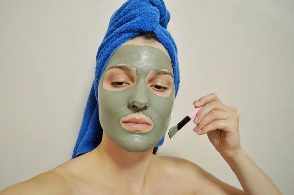 A woman with a brush applies a clay mask to her face with a blue towel on her head