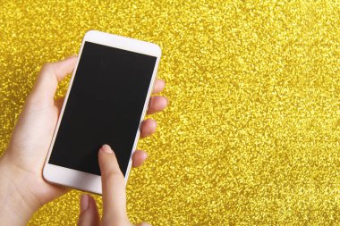 Güzel bir genç kadının eli, parlak sarı bir Noel arka planında siyah ekranlı beyaz bir cep telefonu tutuyor. Boşluğu kopyala.