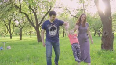 Güzel aile zevk yaz günü parkta: annem ve babam ilk adımlarını yapmak için ona yardım ile yürümeyi öğrenen küçük bebek