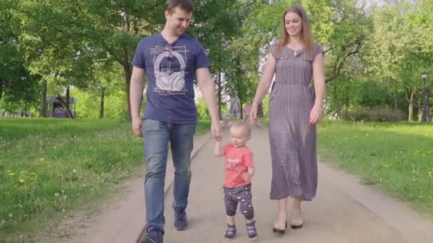 Schöne Familie genießt Sommertag im Park: Kleines Baby lernt mit Mama und Papa laufen und hilft ihm, seine ersten Schritte zu machen — Stockvideo