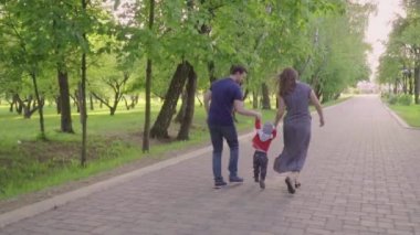 Güzel aile zevk yaz günü parkta: annem ve babam ilk adımlarını yapmak için ona yardım ile yürümeyi öğrenen küçük bebek