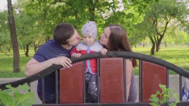 Feliz jóvenes padres compartir beso su lindo bebé chico al aire libre en parque — Vídeos de Stock