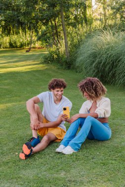 Canlı bir parkta çimlerin üzerinde oturan mutlu genç çift akıllı telefon kullanıyor ve güneşli bir günün tadını çıkarıyor, birlikte gülüyor ve dışarıda anlarımızı paylaşıyorlar.