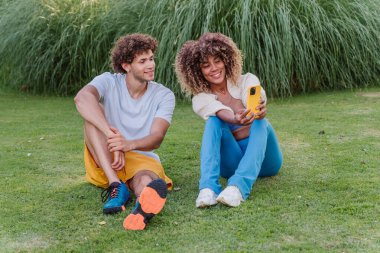 Canlandırıcı bir eğitim seansından sonra çimenlerde oturan genç sportif çoklu etnik çift akıllı telefonlarıyla eğlenceli bir selfie çekiyor, birlikte gülümsüyor ve kaliteli vakit geçiriyorlar.