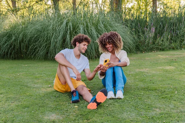 Genç bir adam ve kadın parkta çimlerin üzerinde oturuyor, akıllı bir telefon kullanıyor ve doğayla ve yaz güneşinin sıcaklığıyla çevrili, birlikte sakin bir anın tadını çıkarıyorlar.