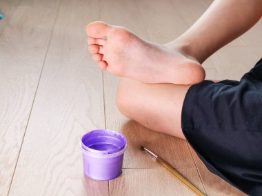 Bir kız boya kutusunun yanında yerde oturuyor ve bacağını boyamak için fırça kullanıyor. Yan görünüm, kapat.