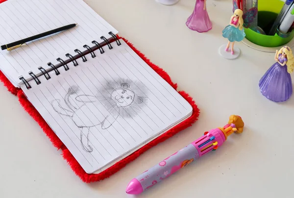 children's notebook with a pattern of a lion, two pens on a white table and dolls in the background, side view close-up.