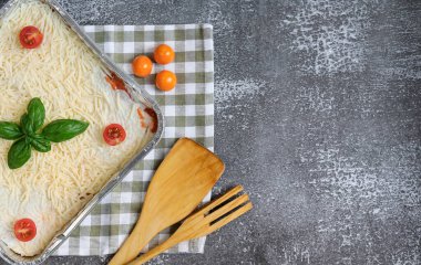 İtalyan lazanyası. Mutfak havlusundaki folyo kabında çiğ lazanya, ahşap bir spatula ve çatalla soldaki gri beton arka planda uzanıyor ve sağdaki yazı için yeri var, üstten yakın çekim..
