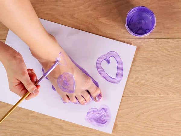 Girl\'s leg and hand with a brush that draws hearts on a white sheet and leg, top view close-up.