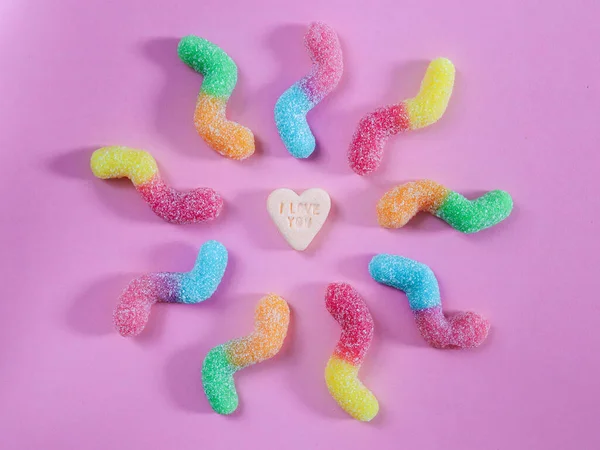 Colored gummy candies in the shape of a zigzag lie in a circle on a lilac background, and in the middle is a candy in the shape of a heart, close-up top view.