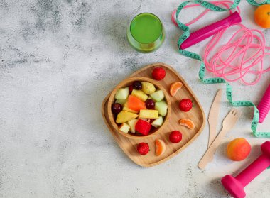 Ahşap tabakta meyve salatası, bardakta sebze püresi ve spor malzemeleri sağ tarafta hafif çimento arka planda ve solda yazı için yer var..