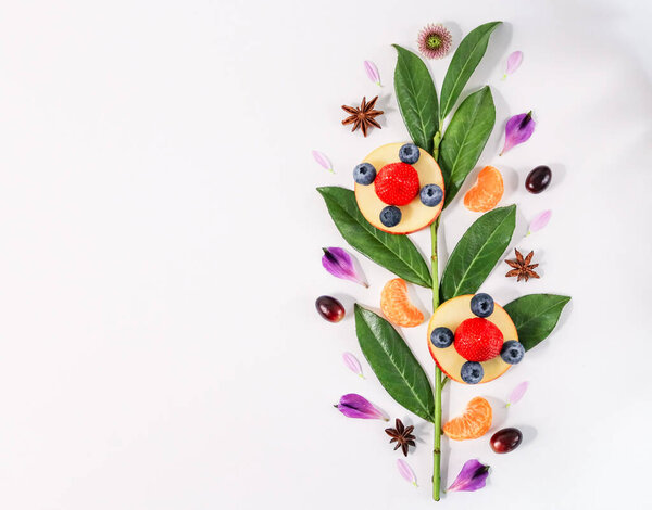 Fruits and berries are laid out in the shape of a flower on a branch with green leaves on the right and with space for text on the left, close-up top view.