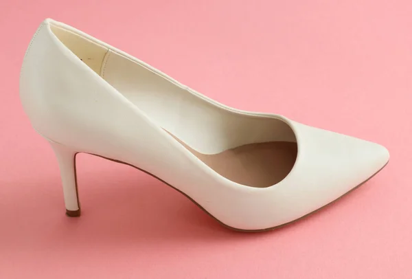 One of white high-heeled shoes stands on a pink background, close-up side view.