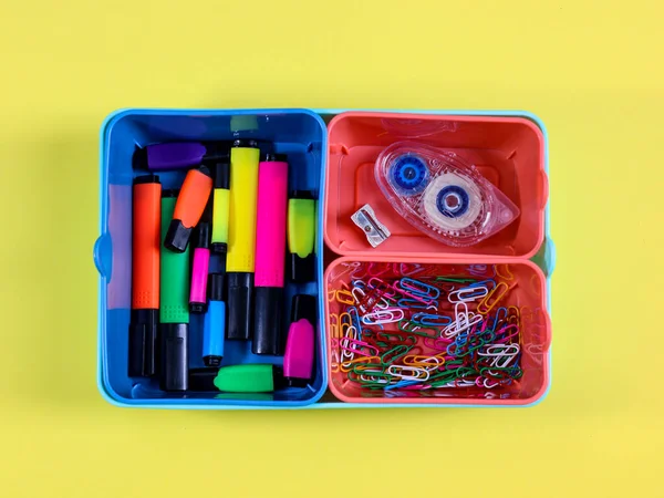 Three plastic colored boxes with markers, paper clips and a marker lie in the middle on a yellow background, close-up top view.