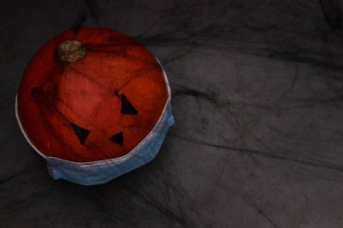 Tıbbi maskeli bir cadılar bayramı balkabağı. Siyah gözlü ve burnu solda. Siyah bir ağda, beton bir arka planda. .