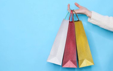 Renkli alışveriş torbaları bir kadının sağ elinde soluk mavi arka planda üç renkli alışveriş torbası ve solda yazı için yer var..