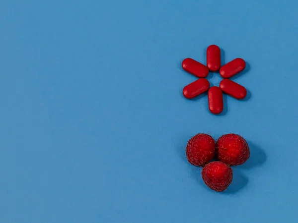 red pills with raspberriesSix red pills and three fresh raspberries lie hardly on a blue background on the right with space for text on the left, close-up top view.
