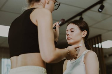 Beyaz tenli genç bir makyöz, sabahın erken saatlerinde güzellik salonunda oturan bir kızın yüzüne kabarık bir fırçayla fondöten tozu sürüyor. Adım adım..