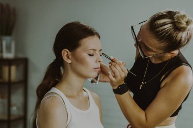 Beyaz tenli genç bir makyaj sanatçısı, sabahın erken saatlerinde bir güzellik salonunda, yakın çekimde bir kızın göz kapaklarına doğal gölgeler sürüyor. Adım adım..