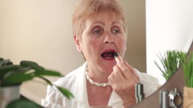 Beyaz bluzlu, güzel, beyaz, yaşlı bir kadın bahar günü yatak odasındaki aynaya bakarak dudaklarını doğal rujla boyuyor. Yan görüntüsü yavaş çekimde. Emeklilik yaşam tarzı kavramı.