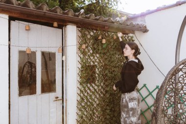 Genç kız yarı yarıya dönmüş ve bağımsız olarak esniyor, bahçedeki bahçe evinin çatısının altına çelenk lambaları asıyor, güneşli bir bahar gününde, seçici bir odak ile kenar manzaralı bir fotoğraf. Çelenk asma zamanı, tatil zamanı..