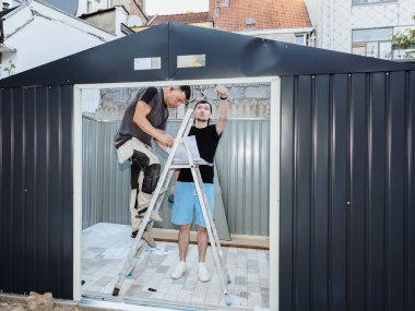 İki beyaz adam merdiven üzerinde konuşuyorlar, bir yaz günü bir evin arka bahçesinde metal bir kulübe yapmak için kağıt talimatlarına bakıyorlar. Metal döşeme ve takım çalışması kavramı.