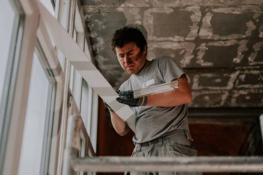 Gri üniformalı yakışıklı beyaz bir adam iskele üzerindeki pencerenin yanındaki bir evde duruyor, eski, kirli bir plastik pencere çerçevesini elinde tutuyor ve tamir edilecek bir cetvelle genişliği ölçüyor, yan görüş. Ev yenileme kavramı