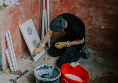 Siyah üniformalı, sarı eldivenli genç beyaz bir adam dizlerinin üzerinde oturuyor ve süngerle yıkıyor. Evin duvarına dayanan plastik bir pencere çerçevesinin bir parçası. Üst tarafı seçici odaklı. Ev yenileme kavramı