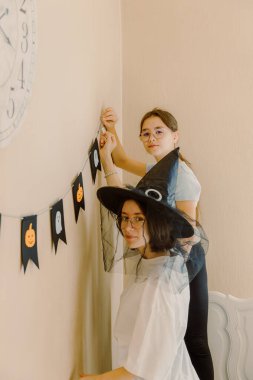 İki beyaz güzel kız kardeşin portresi yatağın üzerinde kameraya bakıyor ve birlikte duvardaki siyah Cadılar Bayramı bayraklarına dekor asıyor, bir sonbahar sabahı yatak odasında tatil için hazırlanıyor,