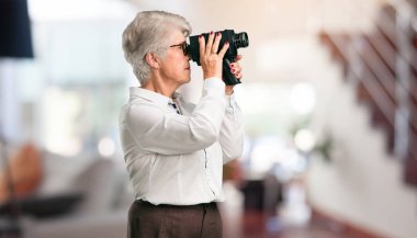 Beautiful senior woman excited and entertained, looking through a film camera, looking for an interesting shot, recording a movie, executive producer at home. clipart