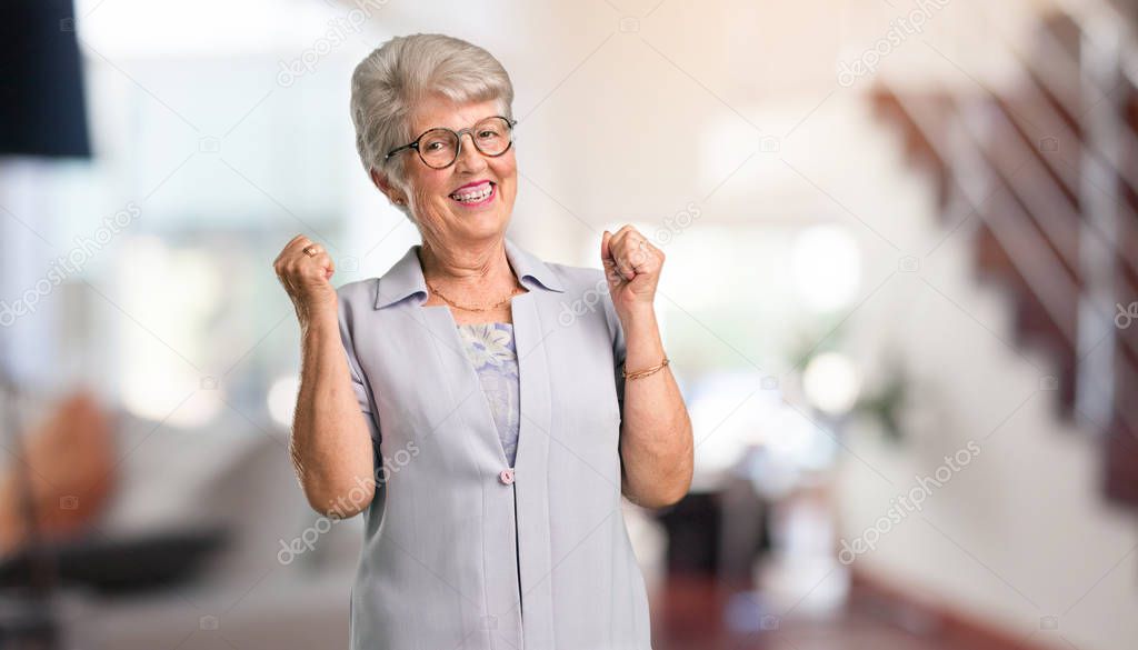 Beautiful senior woman very happy and excited, raising arms, celebrating a victory or success, winning the lottery at home.