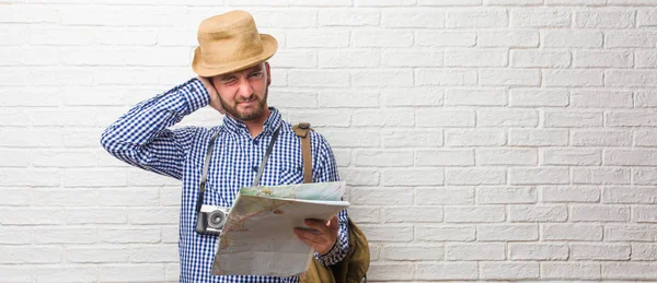 Young traveler man wearing backpack and a vintage camera covering ears with hands, angry and tired of hearing some sound. Holding a map.