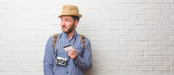 Young traveler man wearing backpack and a vintage camera very scared and afraid, desperate for something, cries of suffering and open eyes, concept of madness. Holding a credit card.