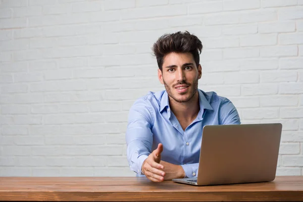 Young business man sitting and working on a laptop reaching out to greet someone or gesturing to help, happy and excited