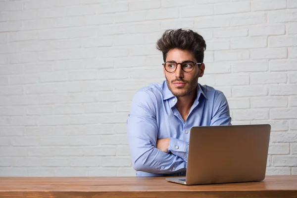 Young business man sitting and working on a laptop doubting and shrugging shoulders, concept of indecision and insecurity, uncertain about something